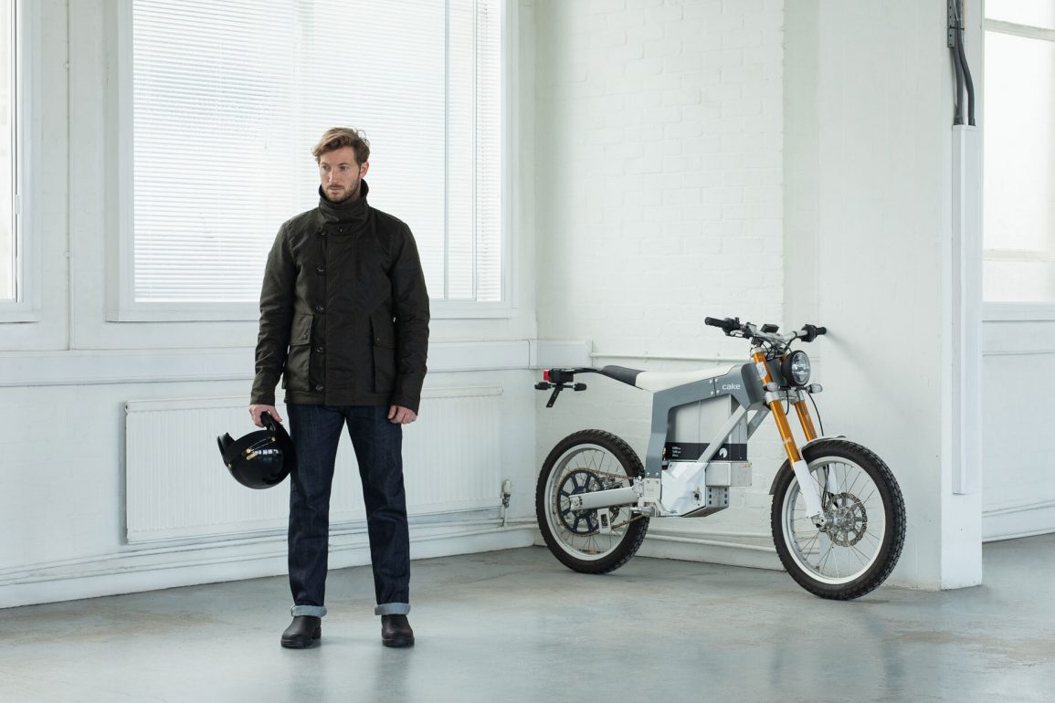 a model wearing the Ashley Watson Armoured Jacket for motorcyclists, next to what appears to be a motor scooter