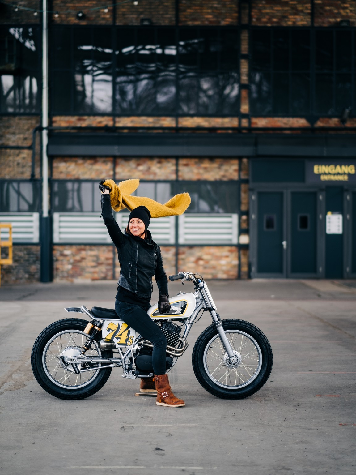 Woman on a Honda Flat Tracker motorcycle waving a yellow scarf and smiling