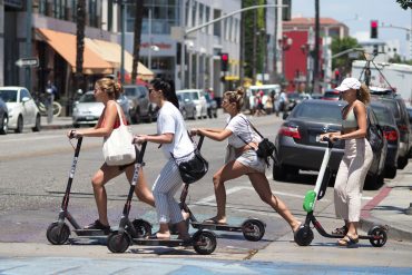 Four women cross a city street with several cars in the background on kickback scooters.